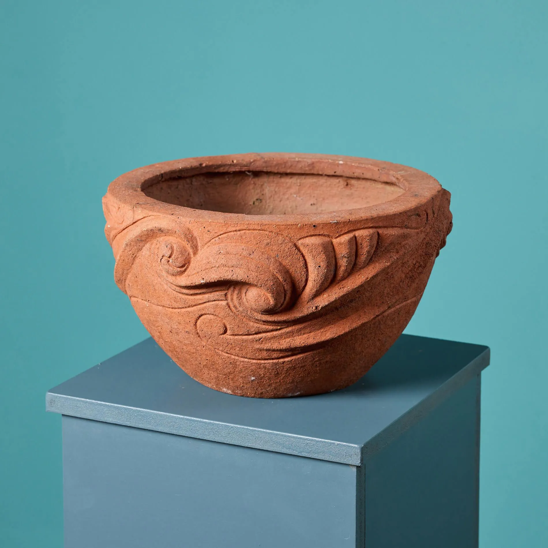 A terracotta pot with intricate swirl and leaf patterns sits atop a blue pedestal against a blue background, reminiscent of designs from Microsoft Montpellier. The pot has a wide, circular opening and an earthy texture.